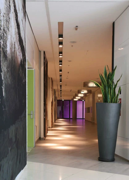 Das neue Foyer im Konferenzbereich besticht durch hochglänzendes Weiss, mit Feinsteinzeug, glatt geputzten Wänden und hochglänzenden Türen. Zur Wirkung kommen Farbe und Materalien durch die Leuchte Prolog 145 in Dolma von Kreon.